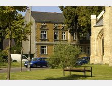 Historisches Wohngebäude mit gelben Klinkersteinen und verzierten Fenstern, im Vordergrund eine Grünfläche mit einem Baum, einem Busch und einer Bank. Rechts im Bild sind Teile eines Gebäudes mit gotischem Spitzbogen aus gelblichem Mauerwerk zu sehen.