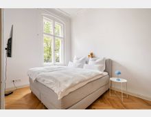Helles Schlafzimmer mit großem Polsterbett und weißer Bettwäsche, neben einem hohen, zweiflügeligen Fenster mit Blick auf Bäume. Weiße Wände mit Stuckdecke, Fischgrätparkett sowie ein kleiner runder Beistelltisch mit blauer Lampe und ein an der Wand montierter Fernseher.