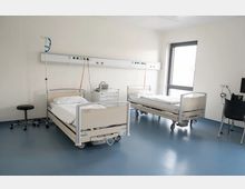 Krankenhauszimmer mit zwei Betten auf Rädern, jeweils mit weißer Bettwäsche. An der Wand befinden sich medizinische Geräteleisten, es gibt ein Fenster mit Lichteinfall, einen schwarzen Hocker sowie einen Schreibtisch mit einem Stuhl. Der Boden ist blau, und die Wände sind weiß.