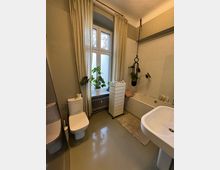 Helles Badezimmer mit beigefarbenem Fliesenboden und einem großen Fenster mit Vorhängen. Im Raum befinden sich eine Toilette, ein Waschbecken, eine Badewanne und ein kleiner Weißschrank; Pflanzen dekorieren die Fensterbank und die Badewanne.