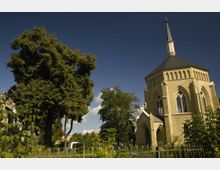 Eine neugotische Kirche mit achteckigem Grundriss, großen Spitzbogenfenstern und einem spitzen Turm, umgeben von Bäumen und einem schmiedeeisernen Zaun. Im Hintergrund sind Teile eines Wohngebiets sichtbar.