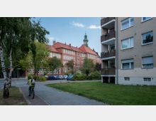 Straßenszene mit einem großen, historischen Backsteingebäude mit rotem Dach und Turmuhr im Hintergrund sowie einem modernen Wohnhaus mit Balkonen rechts im Vordergrund. Grünflächen und Bäume umgeben die Gebäude, während ein Fahrradfahrer auf einem Gehweg vorbeifährt.
