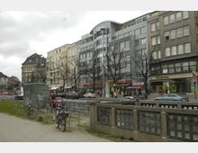 Straßenansicht mit mehrstöckigen Wohn- und Geschäftshäusern, darunter moderne und historische Fassaden. Im Vordergrund stehen Fahrräder an einem Abstellbereich, während Autos und Fußgänger die Straße passieren; Geschäfte wie eine Apotheke und Supermärkte säumen den Gehweg.