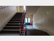 Treppenhaus in einem Innenbereich mit beigen Wänden, dunkelbraunen Stufen und einem roten Geländer. Am unteren Ende ist eine Glastür zu sehen, daneben ein roter Abfalleimer und ein Feuerlöscher. An den Wänden hängen Hinweiszettel und einige Dekorationen.