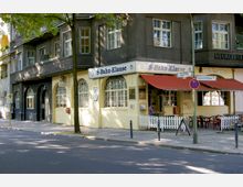 Straßenszene mit einer gelben Fassade eines Restaurants namens "S-Bahn-Klause", das sich in einem Gebäudekomplex mit dunklen Putzwänden und großen Fenstern befindet. Vor dem Lokal stehen rote Markisen, weißer Zaun und Außensitzplätze mit Tischen und Stühlen.