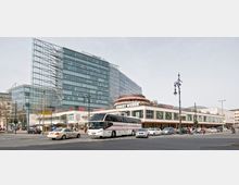 Straßenszene mit einem modernen Glasgebäude im Hintergrund und einem Einkaufszentrum mit Geschäften wie Gerry Weber und H&M im Vordergrund, daneben ein großer Reisebus und mehrere Autos auf der Straße. Die Umgebung zeigt Laternen im historischen Stil und einen belebten Verkehr.