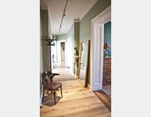 Ein schmaler Flur mit hellgrünen Wänden und einem Holzboden. Auf der linken Seite steht ein Holzstuhl und ein Kleiderständer, rechts ein großer, an die Wand gelehnter Spiegel sowie eine Ablage mit Blumen; im Hintergrund sind geöffnete Türen sichtbar.