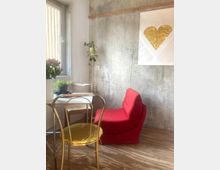 Kleiner Raum mit Betonwand, einem goldfarbenen Stuhl und einem weißen Tisch mit Pflanzen. Rechts steht ein roter Sessel, darüber hängt ein Bild mit einem goldenen Herz auf weißem Hintergrund.