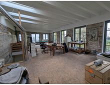 Ein geräumiger Raum mit unverputzten Backsteinwänden und einem Holzdielenboden, eingerichtet mit mehreren Holzmöbeln wie Tischen und Regalen. Die großen Fenster mit schwarzem Rahmen lassen viel Tageslicht herein, während eine sichtbare weiße Balkendecke und ein moderner Metallkaminrohr das industrielle Design unterstreichen.