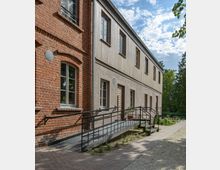 Zweistöckiges Gebäude mit einer Fassade aus rotem Backstein und Holzverkleidung, ergänzt durch einen barrierefreien Zugang mit Rampe und schwarzem Geländer. Im Vordergrund führen ein gepflasterter Weg und ein kleiner Schotterbereich entlang der Gebäudeseite.