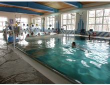 Hallenbad mit einem rechteckigen Becken, in dem Menschen schwimmen und sich entspannen. Große Fenster lassen Tageslicht herein, während blaue und weiße Fliesen das Innere dekorieren; im Hintergrund sind Schwimmhilfen und Sitzgelegenheiten sichtbar.