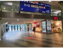 Innenbereich eines Bahnhofs mit breiten Fliesenböden, Glas-Eingangstüren im Hintergrund, und mehreren Geschäften, darunter eine Ditsch-Bäckerei auf der rechten Seite. Über den Geschäften hängen blaue Orientierungsschilder mit Hinweisen zu Bahnsteigen, WC und Ausgängen.