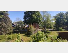 Gartenlandschaft mit vielen Bäumen, Büschen und Rasenflächen, im Hintergrund mehrere Wohnhäuser mit roten und dunklen Dächern, umgeben von weiteren Bäumen. Eine Straße verläuft zwischen den Häusern und den Gärten.