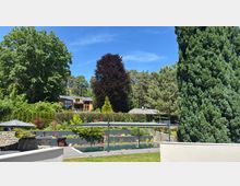 Ein moderner Garten mit einem rechteckigen Pool, umgeben von Steinmauern, Sonnenschirmen und Pflanzenbeeten. Im Hintergrund stehen große Bäume und ein Holzhaus mit Glasfront, eingebettet in eine grüne Landschaft.