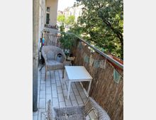 Ein schmaler Balkon mit weiß gefliesten Boden, ausgestattet mit zwei geflochtenen Sesseln und einem kleinen weißen Tisch. Die Brüstung ist mit Bambusmatten verkleidet, darüber hängen dekorative Girlanden; im Hintergrund stehen grüne Bäume und klassische Wohngebäude.