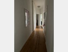 Schmaler Flur in einer Wohnung mit hellen Wänden, Holzboden aus Dielen und einer Deckenlampe; an den Wänden hängen Bilderrahmen, und mehrere Türen führen in angrenzende Räume.