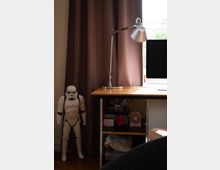 Ein Schreibtisch in einem Innenraum mit einer verstellbaren Tischlampe darauf, flankiert von einer braunen Vorhangkulisse. Unter dem Schreibtisch befinden sich Regalfächer mit verschiedenen Gegenständen, darunter ein kleiner Ventilator und eine rote Figur. Links neben dem Vorhang steht eine große Sturmtruppler-Figur. Im Hintergrund ist teilweise ein Fenster erkennbar.