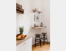 Kleine, modern eingerichtete Küche mit einer hellen Arbeitsplatte, einer Obstschale im Vordergrund und einer Wandbar mit zwei dunklen Hockern. Wandregale mit Dekorationen, ein gerahmtes Bild und Holzboden prägen die minimalistische Gestaltung.