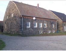 Steingebäude mit Satteldach, errichtet aus unregelmäßigen Feldsteinen, mit mehreren weißen Fenstern und Dachziegeln. Der Boden besteht aus Kopfsteinpflaster, und Satellitenschüsseln sind an der Fassade montiert.