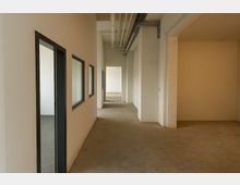 Langer, leerer Flur in einem umgebauten Industrie- oder Bürogebäude mit Betonboden, weißen Backsteinwänden und sichtbaren Deckenleitungen. Links befinden sich schwarze Tür- und Fensterrahmen zu angrenzenden Räumen, rechts öffnet sich ein breiter Durchgang in einen weiteren Raum.