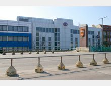 Moderne Bürogebäude mit weißen und blauen Fassadenelementen, gekennzeichnet durch ein C&A-Logo. Im Vordergrund ist eine mehrspurige Straße mit Absperrungen und Geländern erkennbar.