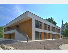 Zweistöckiges modernes Gebäude mit heller Klinkerfassade und vertikalen Holzverkleidungen, einer Außentreppe und großen Fenstern; im Hintergrund ist der Turm einer Kirche zu sehen, umgeben von Bäumen.