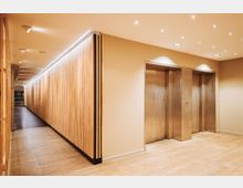 Flur in einem modernen Gebäude mit hellen Holzelementen an der Wand, eingebauten Deckenstrahlern und zwei silbernen Aufzugstüren auf der rechten Seite. Der Boden ist mit hellen Fliesen ausgelegt.