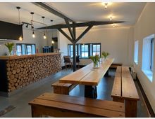 Ein heller Raum mit modern-rustikalem Design, bestehend aus Holzbänken und langen Esstischen. Im Hintergrund befindet sich eine Bar mit einer Verkleidung aus gestapelten Holzscheiten, begleitet von hängenden Lampen und großen Fenstern.