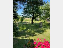 Große, gepflegte Rasenfläche mit schattenspendendem Baum in der Mitte und mehreren verstreut stehenden Metall-Gartentischen und -stühlen. Im Vordergrund blühen pinke Blumen, im Hintergrund sind Felder und einige niedrige Gebäude am Rand des Gartens zu sehen.