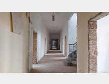 Langer, schmaler Flur in einem Gebäude in Renovierung mit weißen Wänden, freiliegenden Ziegeln um die Türrahmen und einer Treppe mit metallischem Geländer auf der rechten Seite. Der Boden ist teils unvollständig, und die Elektrik an der Decke ist sichtbar.