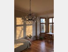 Ein helles Zimmer mit Holzboden, großen Sprossenfenstern und einem Kristallkronleuchter. Der Raum enthält ein Einzelbett, einen Holztisch mit passendem Stuhl und einige Pflanzen, mit Sonnenlicht, das durch die Fenster fällt.