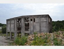 Ein verlassenes, stark beschädigtes Betongebäude mit zerschlagenen Fenstern und freiliegender Struktur, umgeben von überwuchertem Boden und Bauschutt. Im Hintergrund sind Bäume und ein bewölkter Himmel zu sehen.