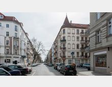 Straßenszene in einer Wohngegend mit mehrstöckigen Altbauten, teils mit Stuckverzierungen und Balkonen. Die Straße ist gesäumt von geparkten Autos, Bäumen ohne Laub und weist ein kleines Geschäft im Erdgeschoss eines Gebäudes auf.