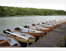 Holzboote sind an einem Steg aus dunklem Holz vertäut, die sich in einem ruhigen See befinden, umgeben von dichtem Wald im Hintergrund. Der Himmel ist leicht bewölkt, und die Szene wirkt ruhig.