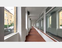 Ein überdachter Korridor mit roten Fliesenböden und weißen Wänden, rechts begrenzt durch große Glasfenster, die in einen Innenraum blicken, und links mit offenen Bögen, die den Blick auf eine Straße und beige Gebäude ermöglichen.