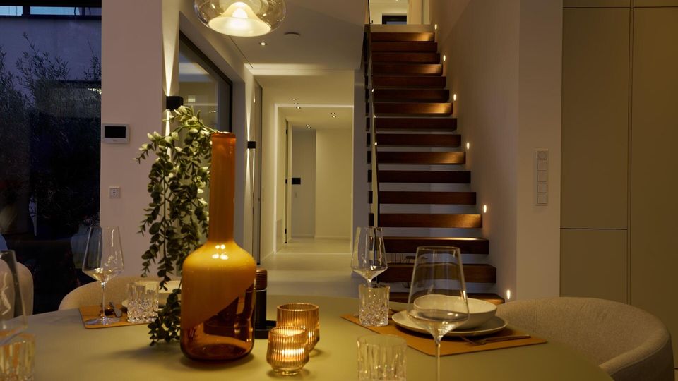 Moderner Wohn-Essbereich mit gedecktem Esstisch im Vordergrund, Glas-Pendelleuchte und dekorativen Vasen sowie Kerzen. Im Hintergrund führt eine offene Holztreppe mit integrierter Stufenbeleuchtung in ein Obergeschoss, daneben ein heller Flur und große Fensterfront.