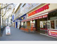 Straßenszene mit einer Reihe von Geschäften, darunter ein „Dunkin’ Donuts“ und ein „Tchibo“-Laden. Vor dem „Dunkin’ Donuts“ befinden sich orangefarbene Außenstühle und Tische; links ist ein Bürgersteig mit kahlen Bäumen und Passanten.