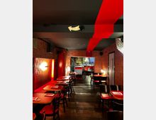 Ein stilvoll eingerichtetes Restaurant mit roten und schwarzen Akzenten, dunklem Holzboden und dekorativen Mustertapeten. Im Hintergrund befindet sich eine Bar mit Barhockern und beleuchteten Flaschenregalen.