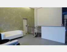 Minimalistisch eingerichteter Innenraum mit einer weißen Couch links, einer kleinen weißen Theke rechts und einer Tür im Hintergrund. Eine Wendeltreppe mit Geländer führt von der Theke aus nach unten.