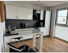 Moderne Küche mit weißen Hochglanz-Schränken, schwarzer Arbeitsplatte und integrierten Elektrogeräten. Im Vordergrund steht ein hoher Tisch mit Glasoberfläche und Barhocker, während ein Fenster rechts den Raum mit Tageslicht beleuchtet.