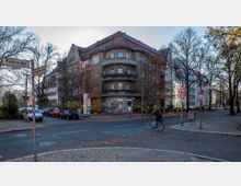 Straßenszene in einer Wohngegend mit einem dreistöckigen Eckgebäude im Hintergrund, geschmückt mit Blumenkästen an den Balkonen. Im Vordergrund eine Kreuzung mit Straßenschildern ("Parkstraße" und "Amalienstraße"), Verkehrszeichen und einem Radfahrer; links sind parkende Autos sowie herbstliche Bäume sichtbar.