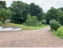 Gepflasterter Weg in einer parkähnlichen Umgebung mit Grünflächen, Blumenbeeten und Bäumen im Hintergrund. Rechts säumt ein Busch mit lilafarbenen Blüten den Weg, der leicht geschwungen verläuft.