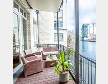 Wohnbalkon mit Holzboden, ausgestattet mit Sitzmöbeln aus Polyrattan und einem kleinen Beistelltisch, sowie einer Topfpflanze. Der Balkon bietet Ausblick auf einen Fluss und moderne Gebäude im Hintergrund.
