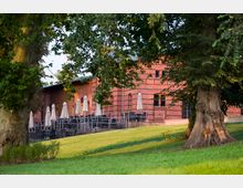 Rote Backsteinfassade eines Gebäudes mit einer großzügigen Terrasse, auf der Tische, Stühle und geschlossene Sonnenschirme stehen, umgeben von Grünflächen und großen Bäumen.