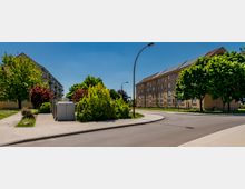 Kurvenreiche Wohnstraße in einem Siedlungsgebiet, gesäumt von mehrstöckigen Wohnblöcken mit Satteldächern. Breite Gehwege, Straßenlaternen sowie dichte Bäume und Sträucher prägen das Straßenbild.