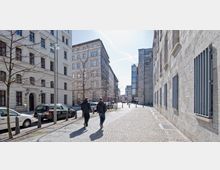 Straßenszene in einer Stadt mit Wohn- und Bürogebäuden aus Steinfassaden; auf der linken Straßenseite parken Autos, während auf der rechten ein Gehweg aus Kopfsteinpflaster verläuft. Zwei Personen gehen auf dem Gehweg, und kahle Bäume stehen entlang der Straße.