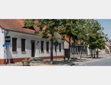 Straßenszene mit einer Reihe von Wohnhäusern mit roten Ziegeldächern und hellen Fassaden, flankiert von Bäumen auf einem gepflasterten Gehweg. Im Vordergrund ist ein Straßenschild mit der Aufschrift „Berliner Straße“ und ein kleines Restaurant-Schild zu sehen.