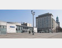 Offener Platz mit einem modernen Gebäude mit der Aufschrift „KOSMOS“ links, gekennzeichnet durch eine Glasfassade und eine Wand mit Mosaiksteinen. Rechts befindet sich ein großes historisches Wohngebäude im Stil des Sozialistischen Klassizismus, im Hintergrund ein Turm mit grüner Kuppel.