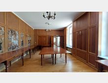 Holzvertäfelter Raum mit rechteckigen Tischen auf Holzfußboden. Die Wände sind mit verzierten Paneelen und Wandgemälden dekoriert, ein Kronleuchter hängt von der Decke, und eine Tür sowie große Fenster lassen Licht herein.