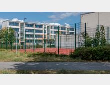 Mehrstöckiges modernes Schulgebäude mit großen Fenstern im Hintergrund, davor ein eingezäunter roter Sportplatz mit umliegenden Grünflächen und einem schmalen Gehweg im Vordergrund.